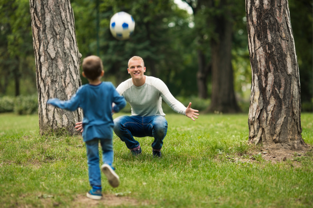 Smiling dad opens his arms to catch soccer ball kicked by his son. 